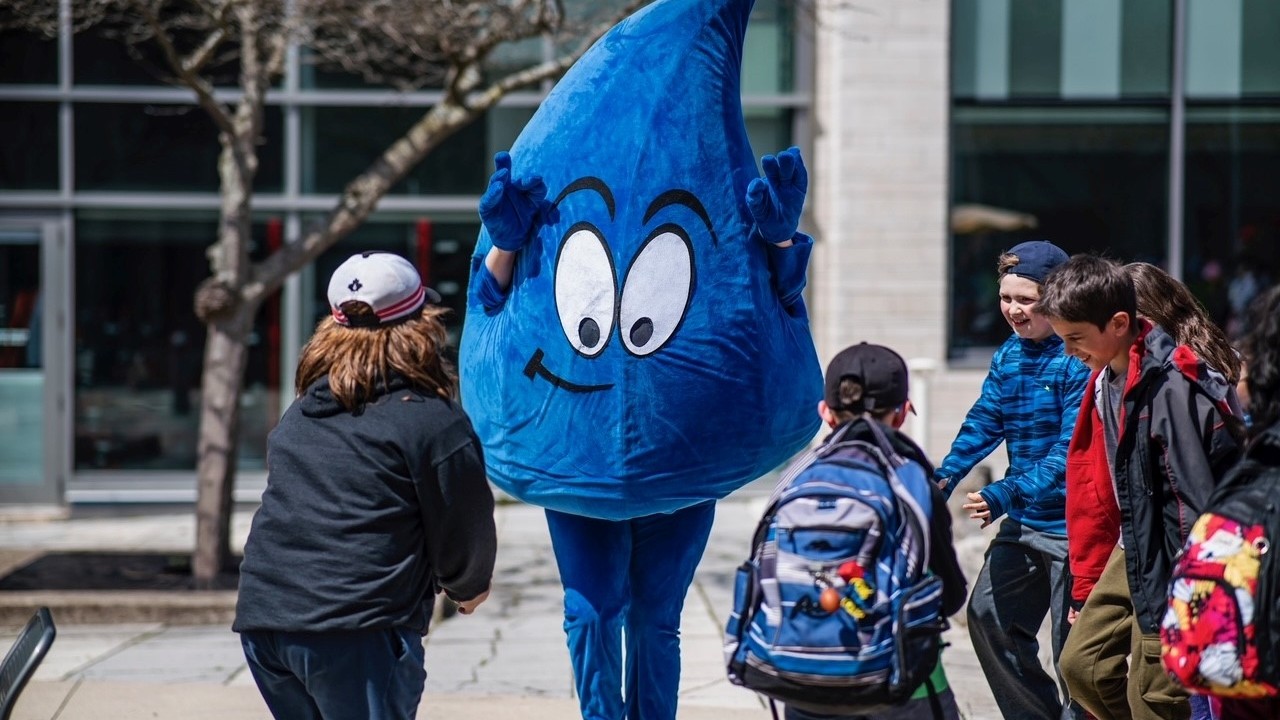 A waterdrop mascot waves at a group of young children.